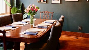 Une salle à manger avec une table en bois dressée pour quatre personnes, entourée de chaises rembourrées. Un lustre est suspendu au plafond et les murs sont ornés d’œuvres d’art encadrées.