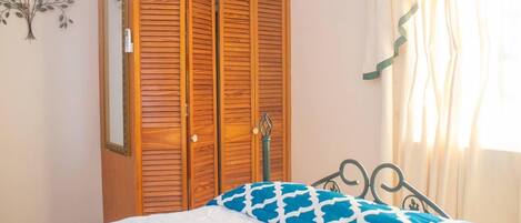 A bedroom with a wooden wardrobe, a bed with a patterned blanket, a wall-mounted light fixture, and a window with sheer curtains.