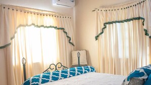 A bedroom with two beds, a window with curtains, a bedside table, and an air conditioning unit. - Lovely 2-bedroom Apt, Sleeps 3, Warm & Cosy (Potters Village)