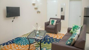 A living room with a sofa, armchair, glass coffee table, and a flat-screen TV mounted on the wall. - Lovely 2-bedroom Apt, Sleeps 3, Warm & Cosy (Potters Village)