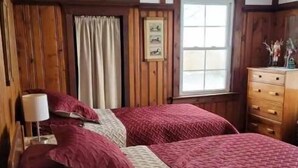 A bedroom with a wooden ceiling, a bed with red and beige bedding, a nightstand with a lamp, a window with curtains, and a dresser with decorative items.
