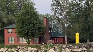 A red house with a chimney, surrounded by trees and a body of water.