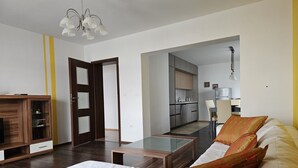 Uma sala de estar moderna com sofá, mesa de centro e armário de madeira. Cozinha visível ao fundo, com armários e área de jantar.
