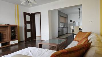A modern living room with a sofa, coffee table, and wooden cabinet. There is a kitchen in the background with cabinets and a dining area.