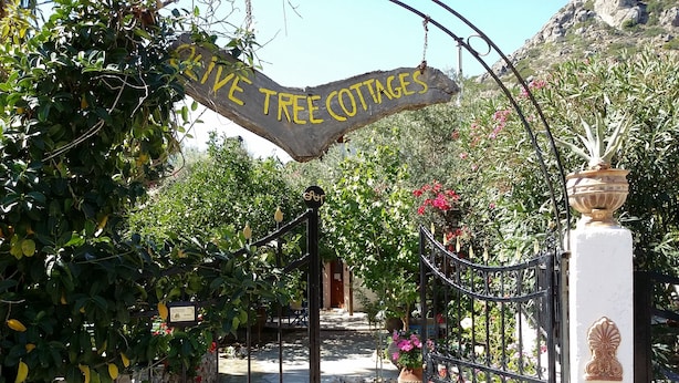 A wrought iron gate with a sign reading "Live Tree Cottages" leading to a pathway surrounded by lush greenery and flowering plants. - Stunning 2-bedrooms Private Cottage in Palaiochora (Kantanos-Selino)