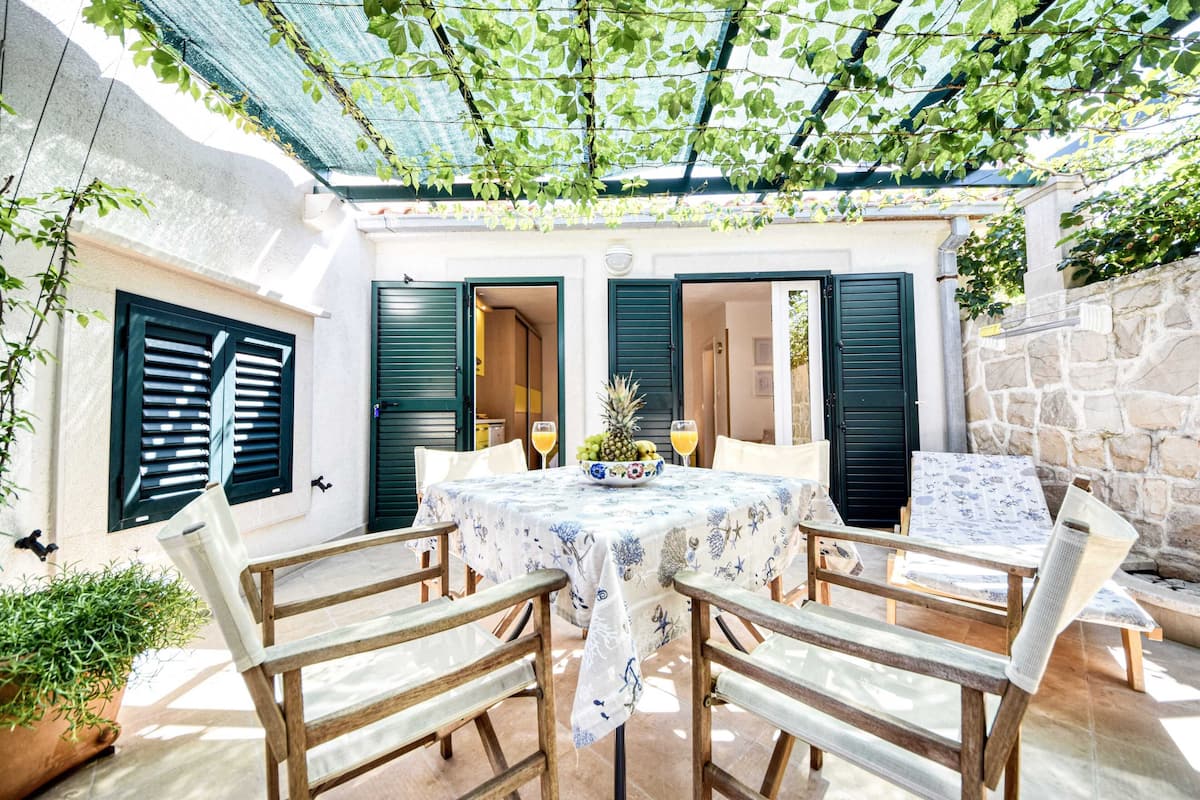 An outdoor dining area with a table set for two, surrounded by wooden chairs, under a pergola with green leaves.