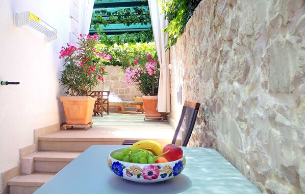 A table with a fruit bowl, a chair, and a potted plant in a sunlit outdoor area with a stone wall and steps leading to a wooden deck. - Apartments Gracin (Selca)