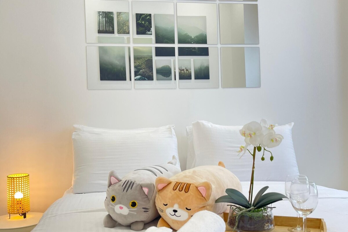 A neatly made bed with white bedding, a small potted plant, a glass of water, and a tray with towels and a cat-shaped pillow.