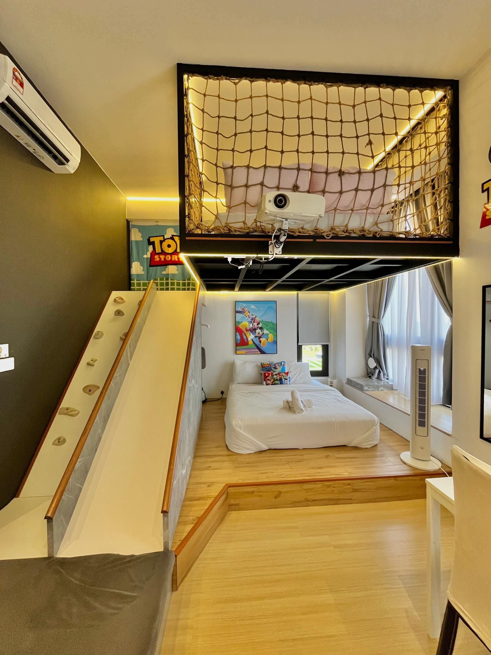 A hotel room with a bed, a desk, a window, a ceiling-mounted net, and a wall-mounted air conditioner.