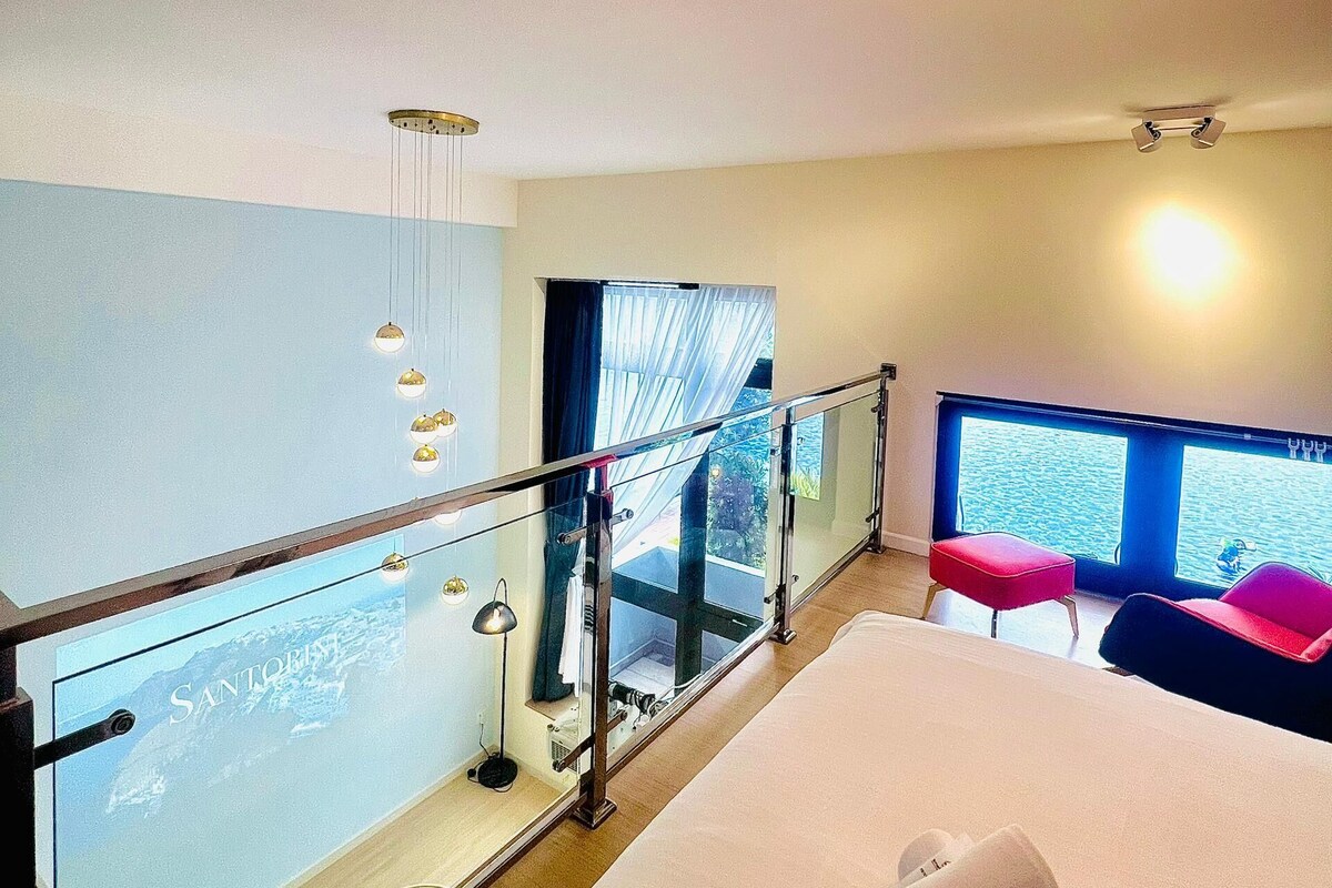 A hotel room with a large bed, a balcony with a glass railing, and a view of the sea.