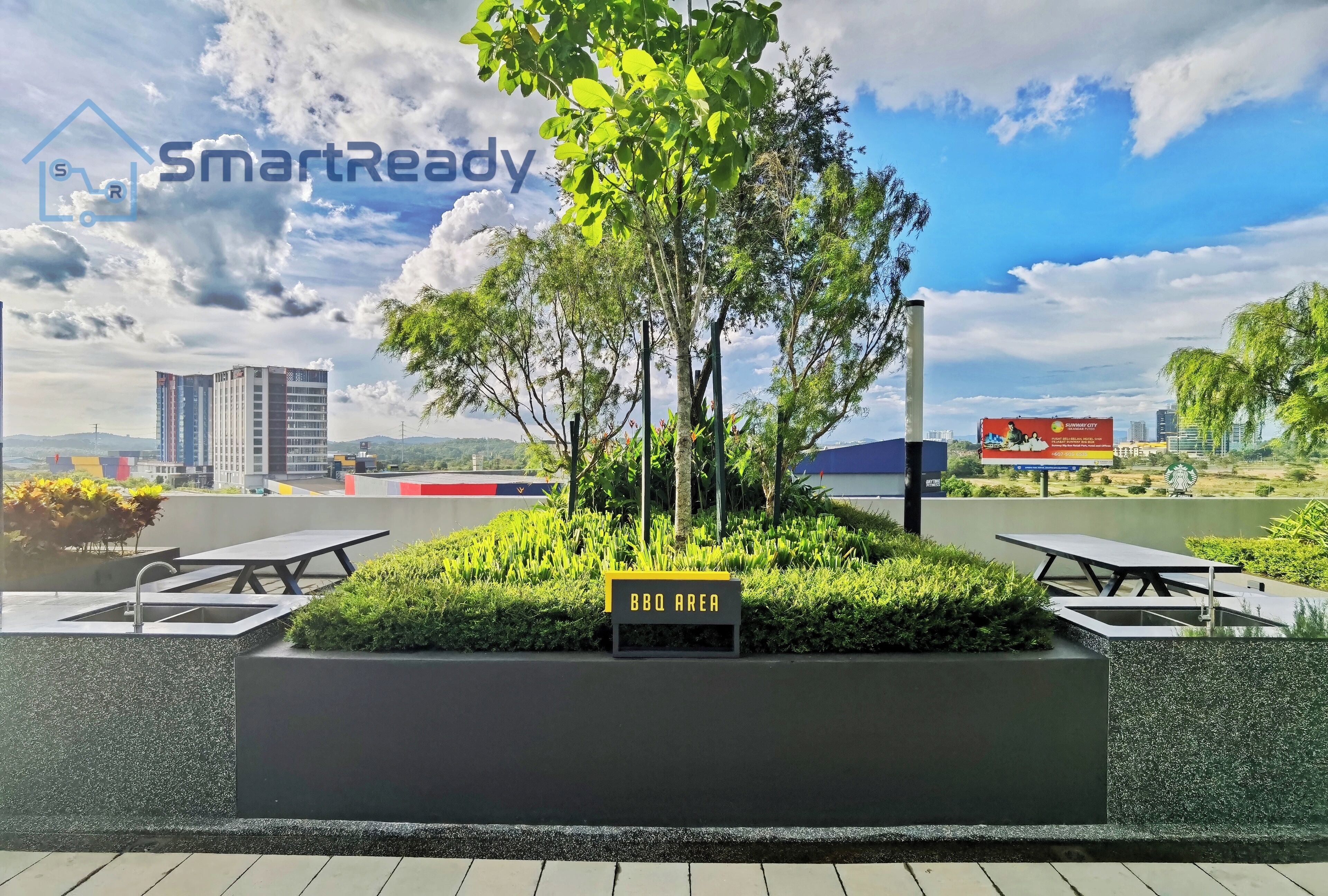 A rooftop BBQ area with a sign, surrounded by greenery and a cityscape in the background.
