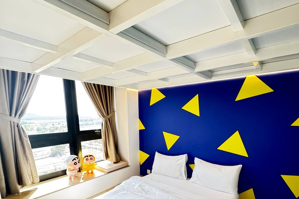 A modern hotel room with a large bed, a view of the city, and a unique blue wall with yellow triangles.