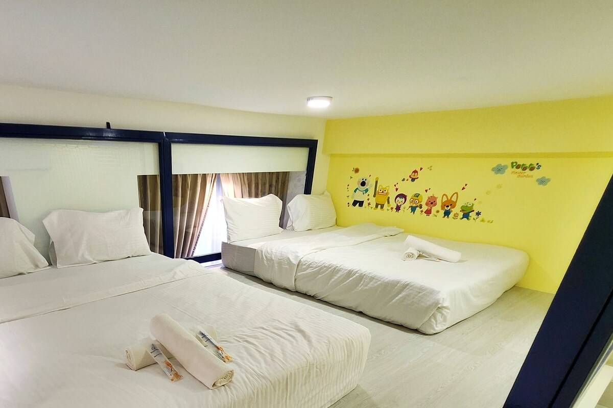 A hotel room with two beds, a large window with curtains, a yellow wall with a children's mural, and a ceiling light.