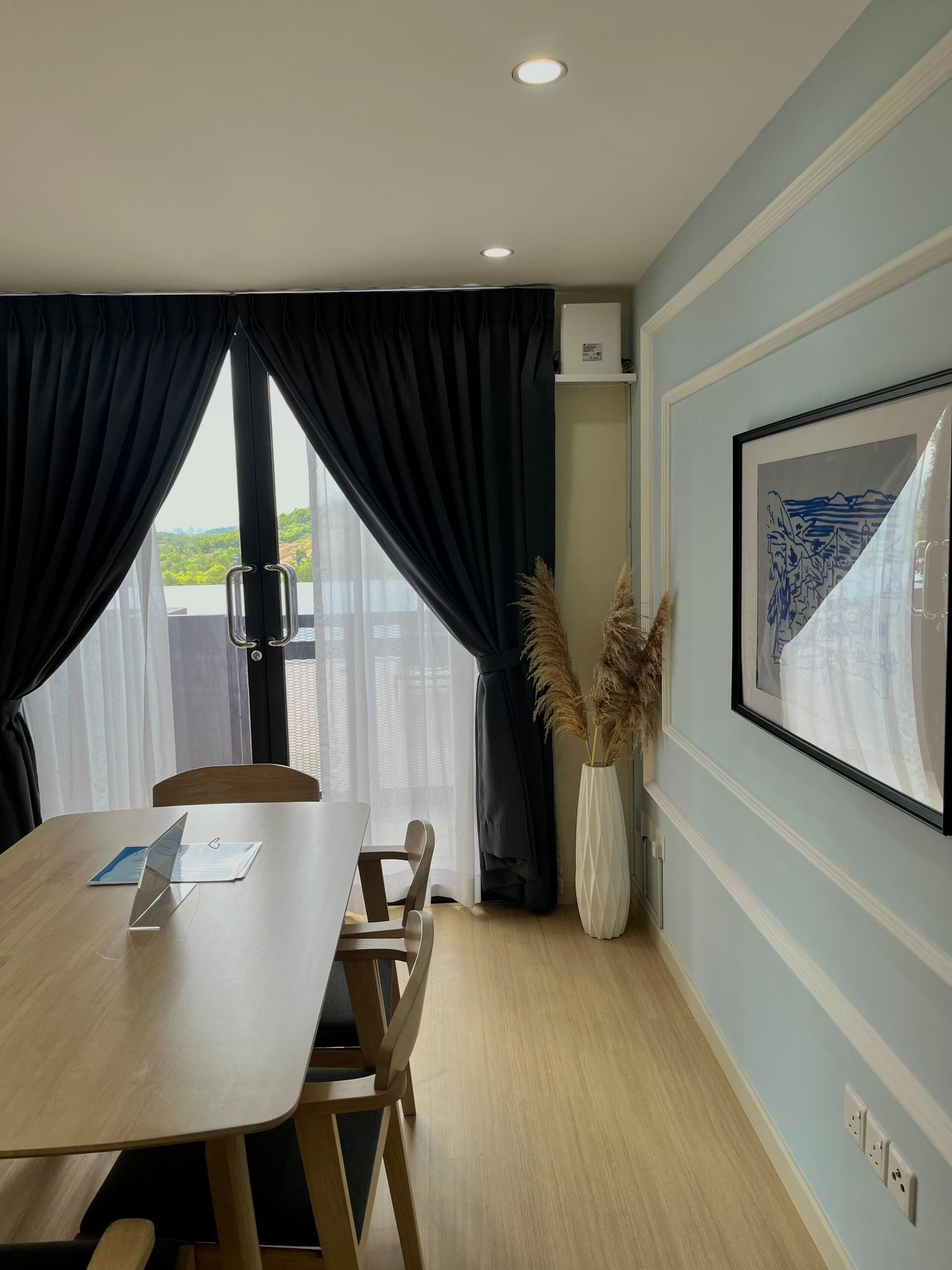 A modern room with a wooden dining table, chairs, a large window with curtains, a framed picture on the wall, and a vase with pampas grass.