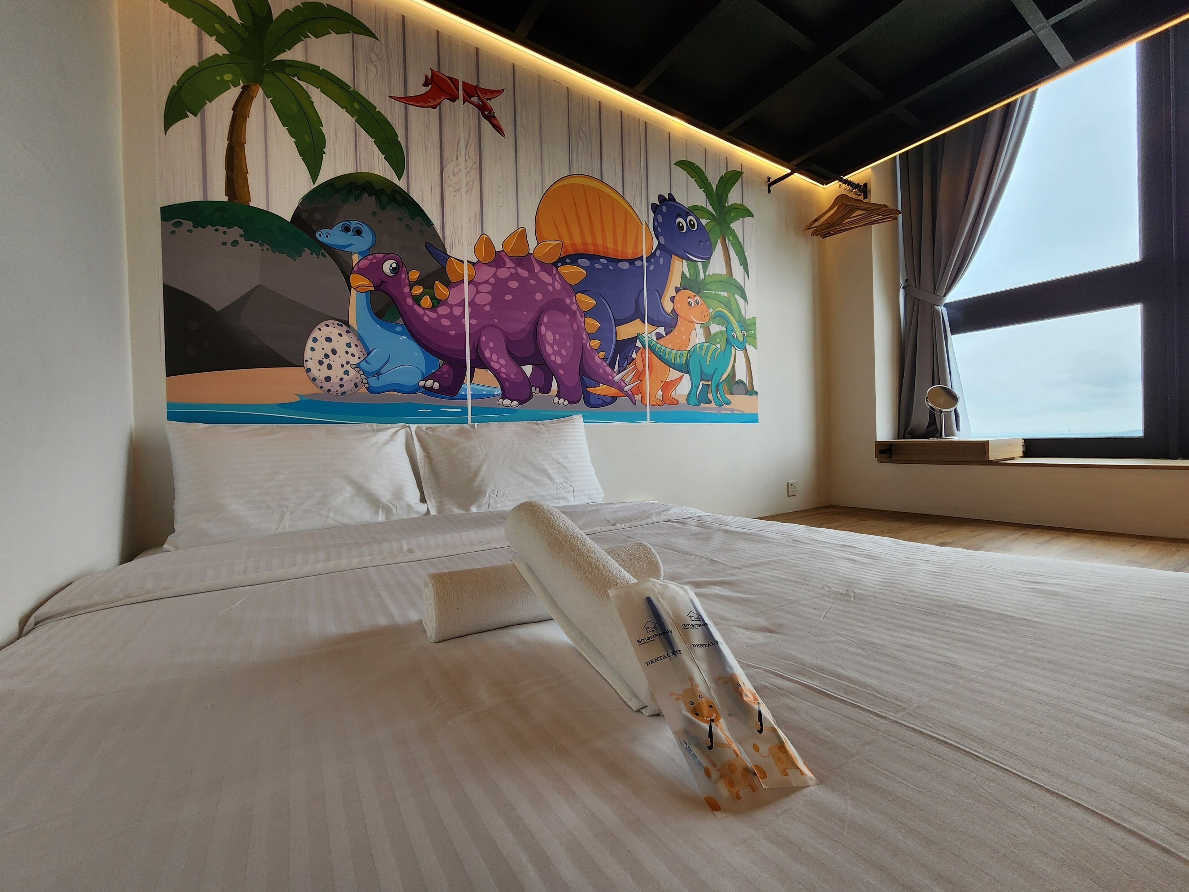 Ein Schlafzimmer mit Bett, weißer Bettwäsche, Wandgemälde mit Dinosauriern, einem Fenster mit Vorhängen und einem Nachttisch mit Lampe.
