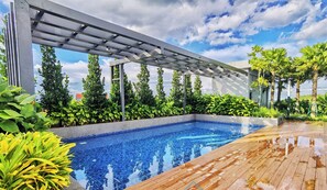 A swimming pool with a wooden deck, surrounded by lush greenery and a pergola structure.