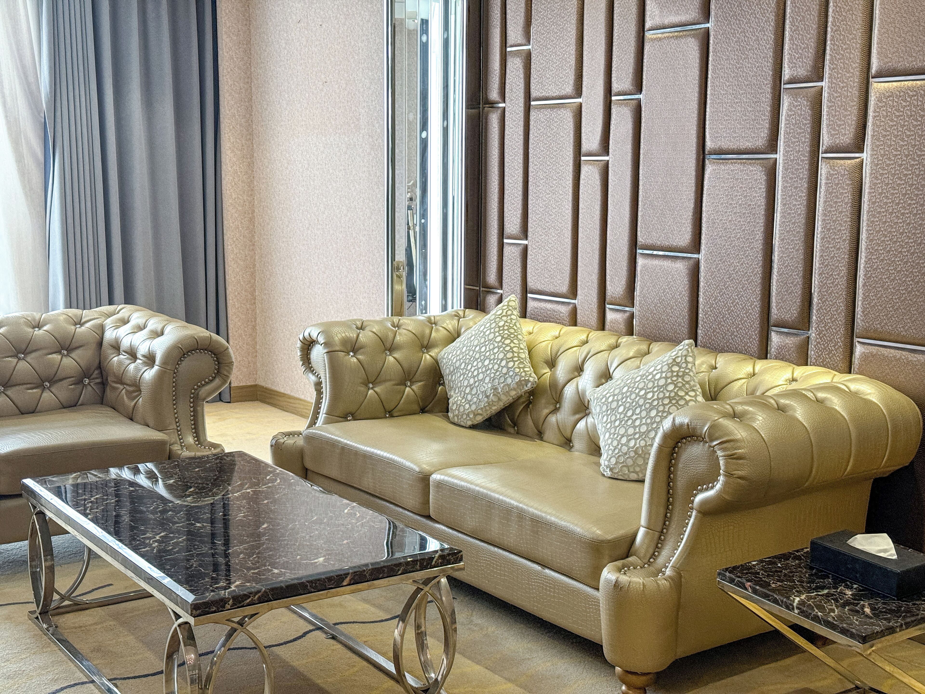 A luxurious living room with a beige leather sofa, two matching armchairs, a marble coffee table, and a decorative wall panel.