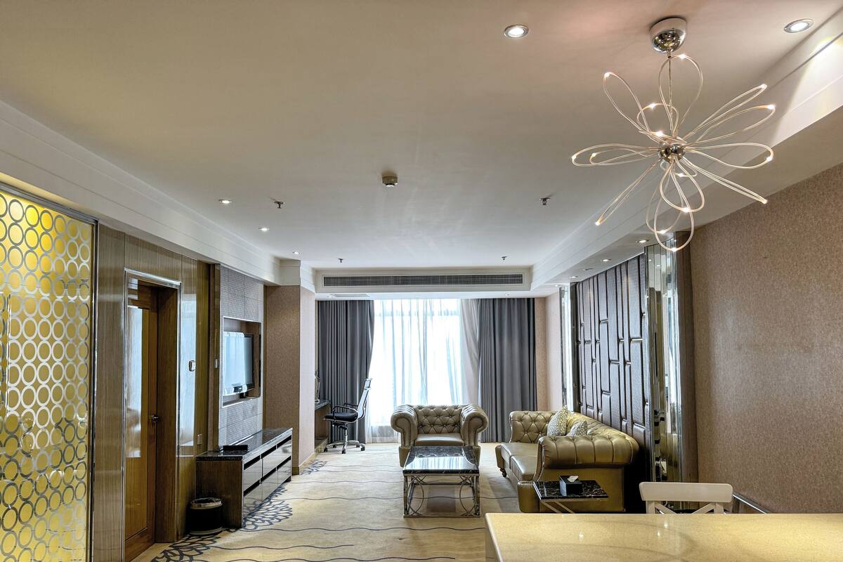 A modern hotel room with a seating area featuring a sofa, armchairs, and a coffee table. There is a flat-screen TV mounted on the wall, a ceiling fan, and a decorative light fixture.