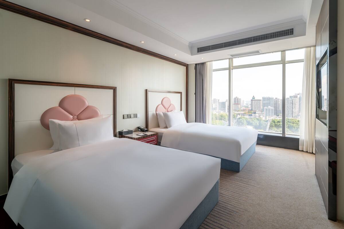 A hotel room with two beds, a large window with a city view, and a flat-screen TV.