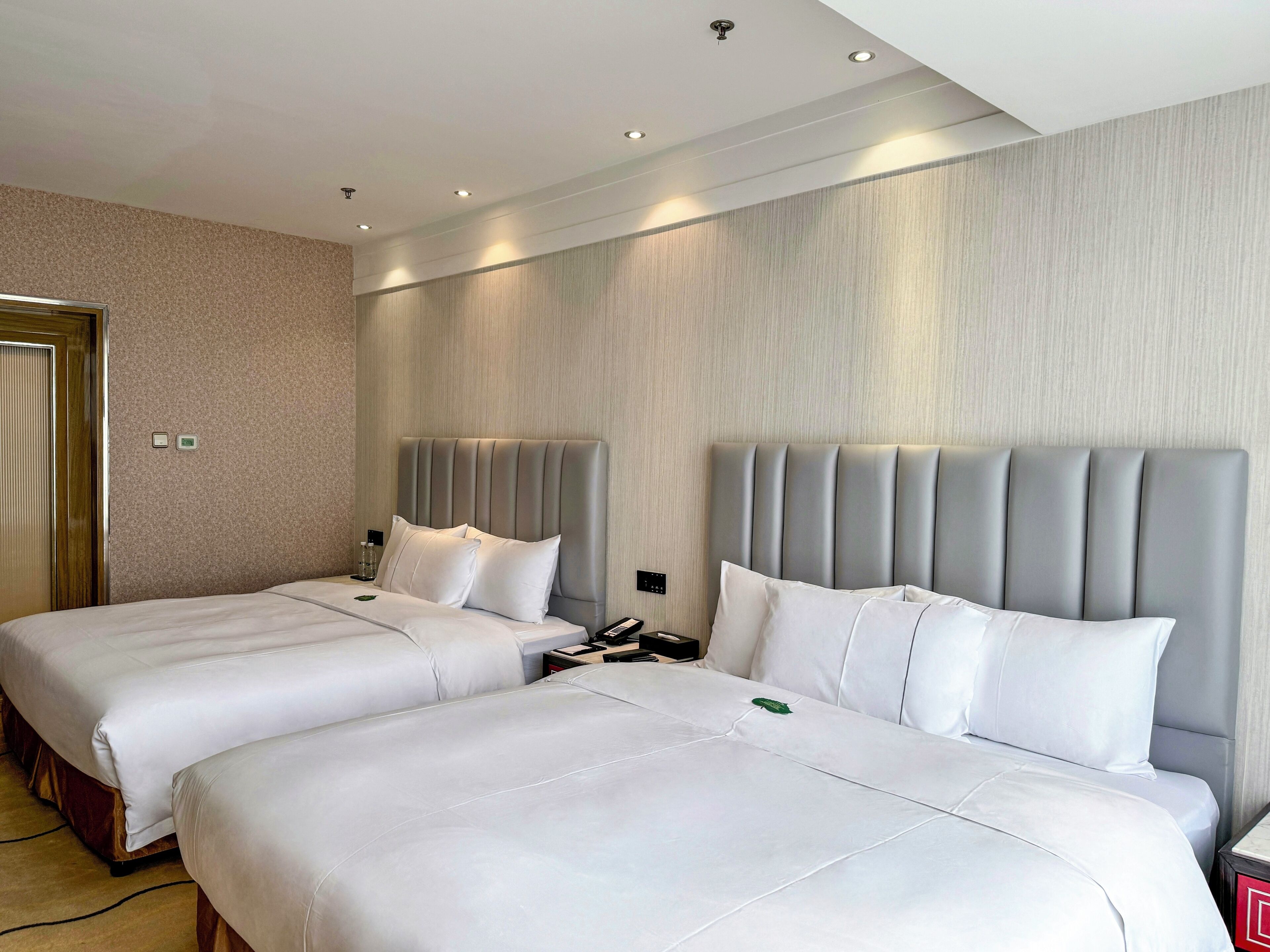 A hotel room with two beds, a wooden door, a bedside table with a phone, and a wall-mounted control panel.