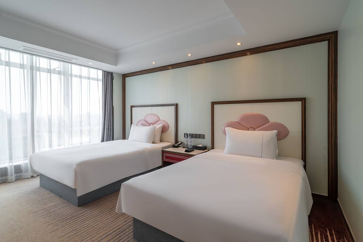A hotel room with two beds, a nightstand, and a large window with sheer curtains.
