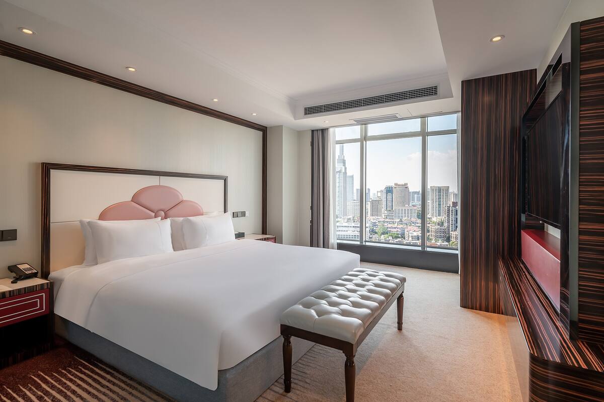 A modern hotel room with a large bed, a bench, a TV, and a city view.
