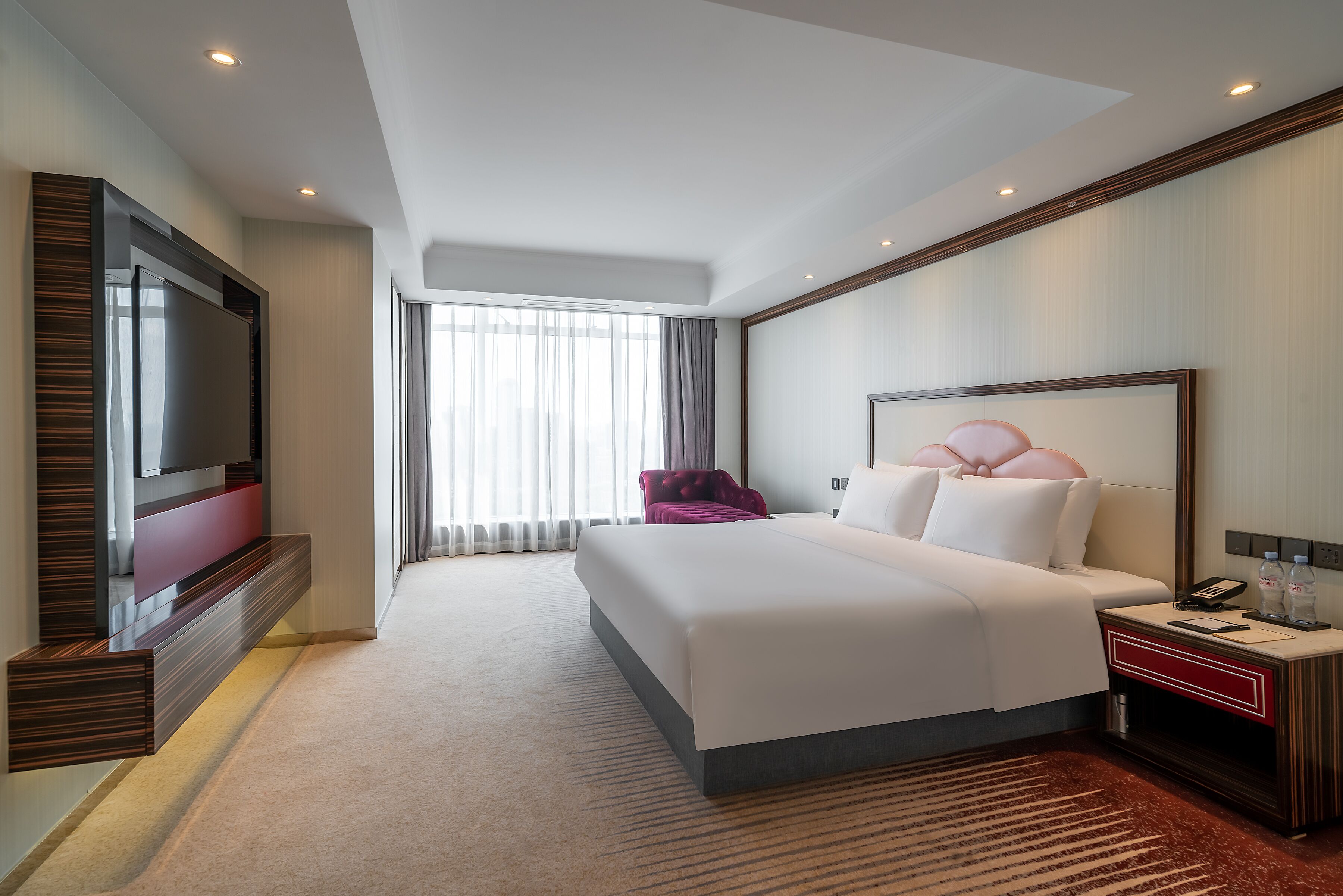 A modern hotel room with a large bed, a TV mounted on the wall, a bedside table with a phone and bottles, and a window with curtains.