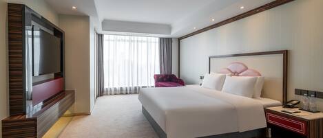 Una habitación de hotel moderna con una cama grande, un televisor en la pared, una mesita de noche con un teléfono y botellas, y una ventana con cortinas.