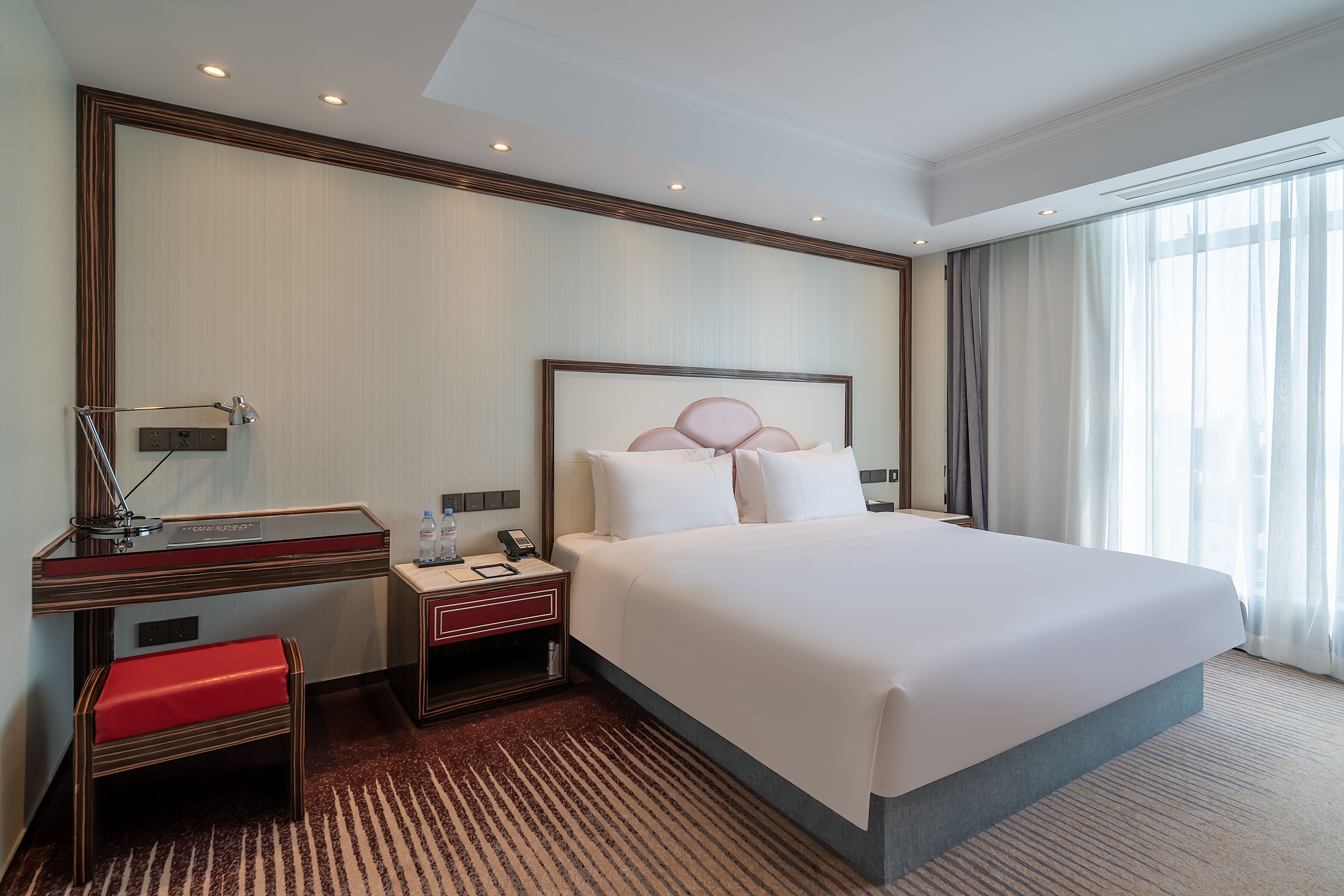Quarto de hotel com uma cama grande, uma escrivaninha com luminária, um banco e vista para o exterior pela janela.