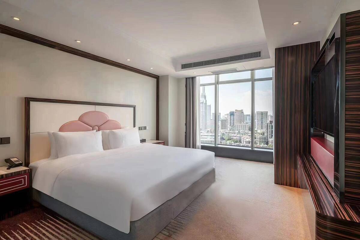 A modern hotel room with a large bed, a city view, a TV, and a desk with a phone.