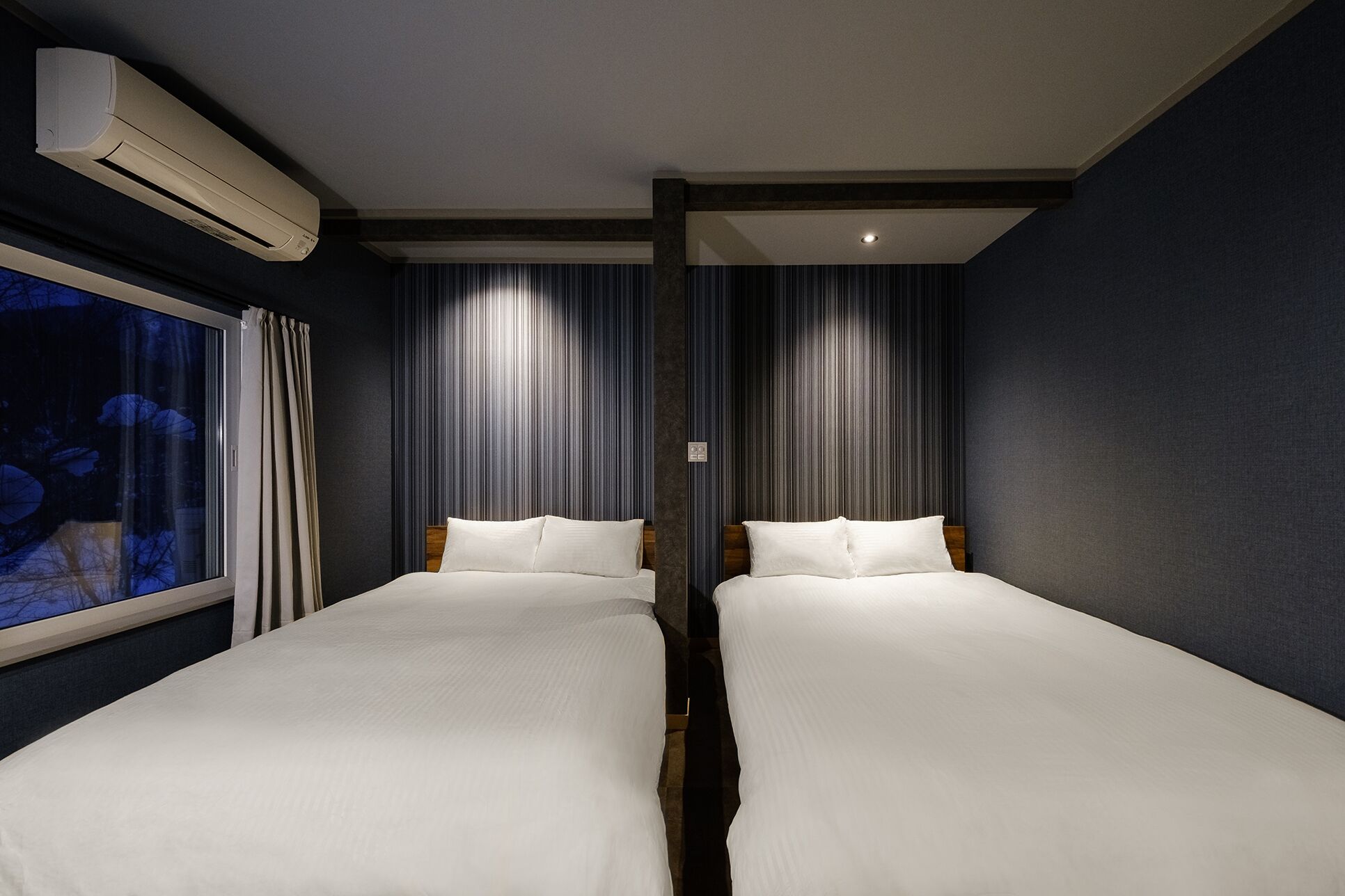 A hotel room with two single beds, a large window with curtains, and an air conditioning unit on the wall.