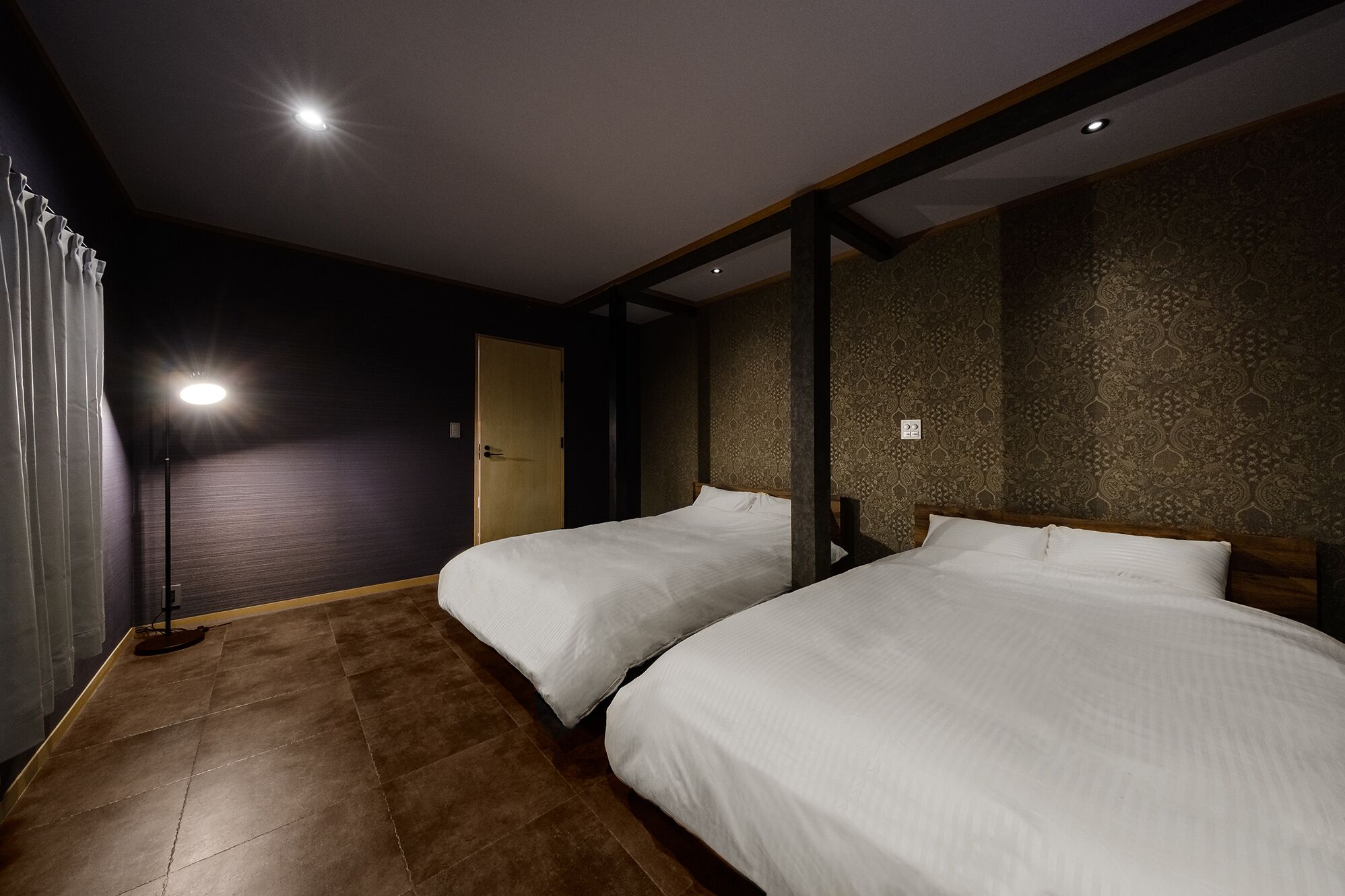 Habitación de hotel con dos camas, piso de madera, pared oscura con papel tapiz estampado y una lámpara en el techo.