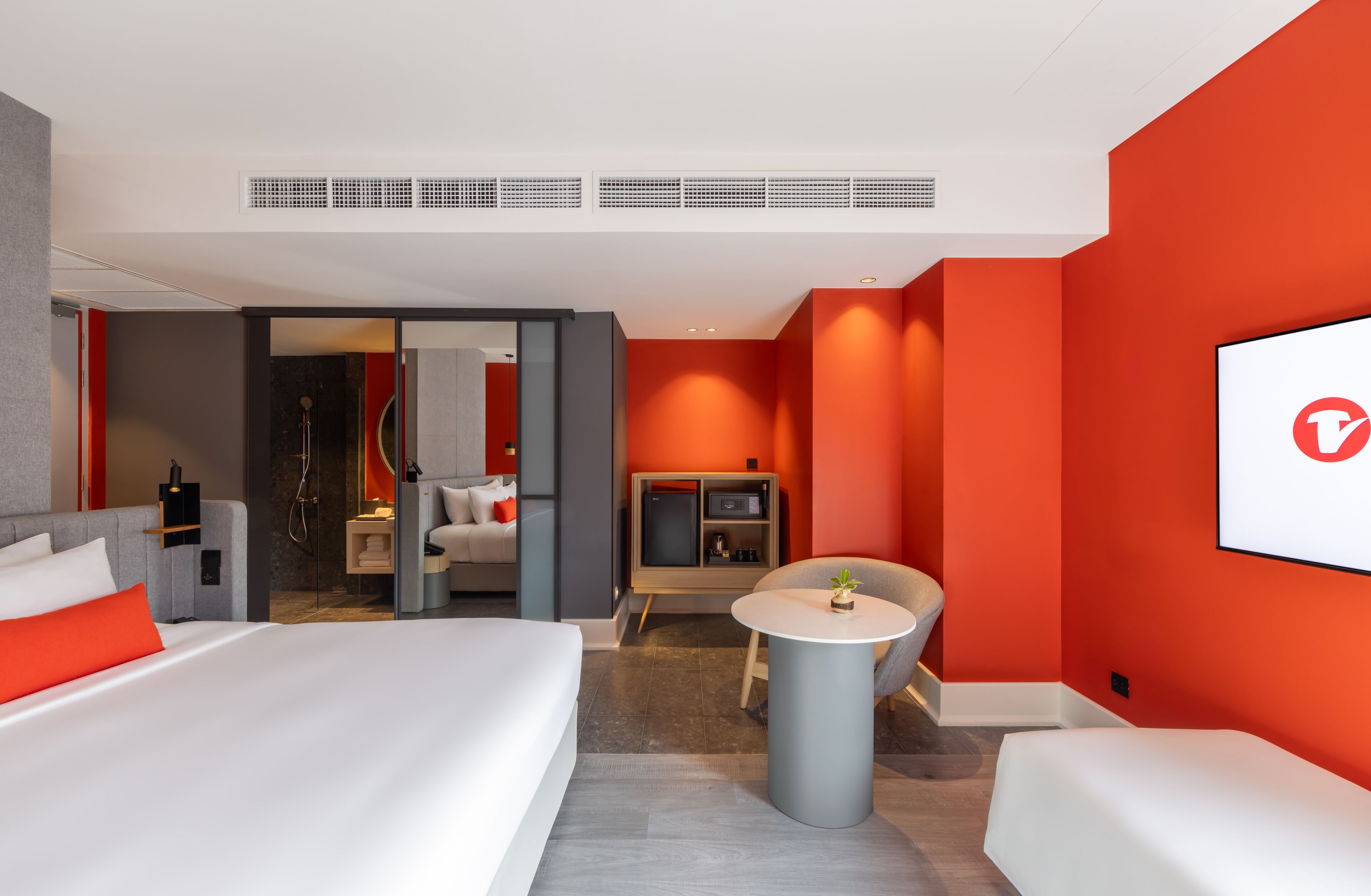 A modern hotel room with a large bed, a desk, a chair, and a small table. There is a TV mounted on the wall and a window with a view.