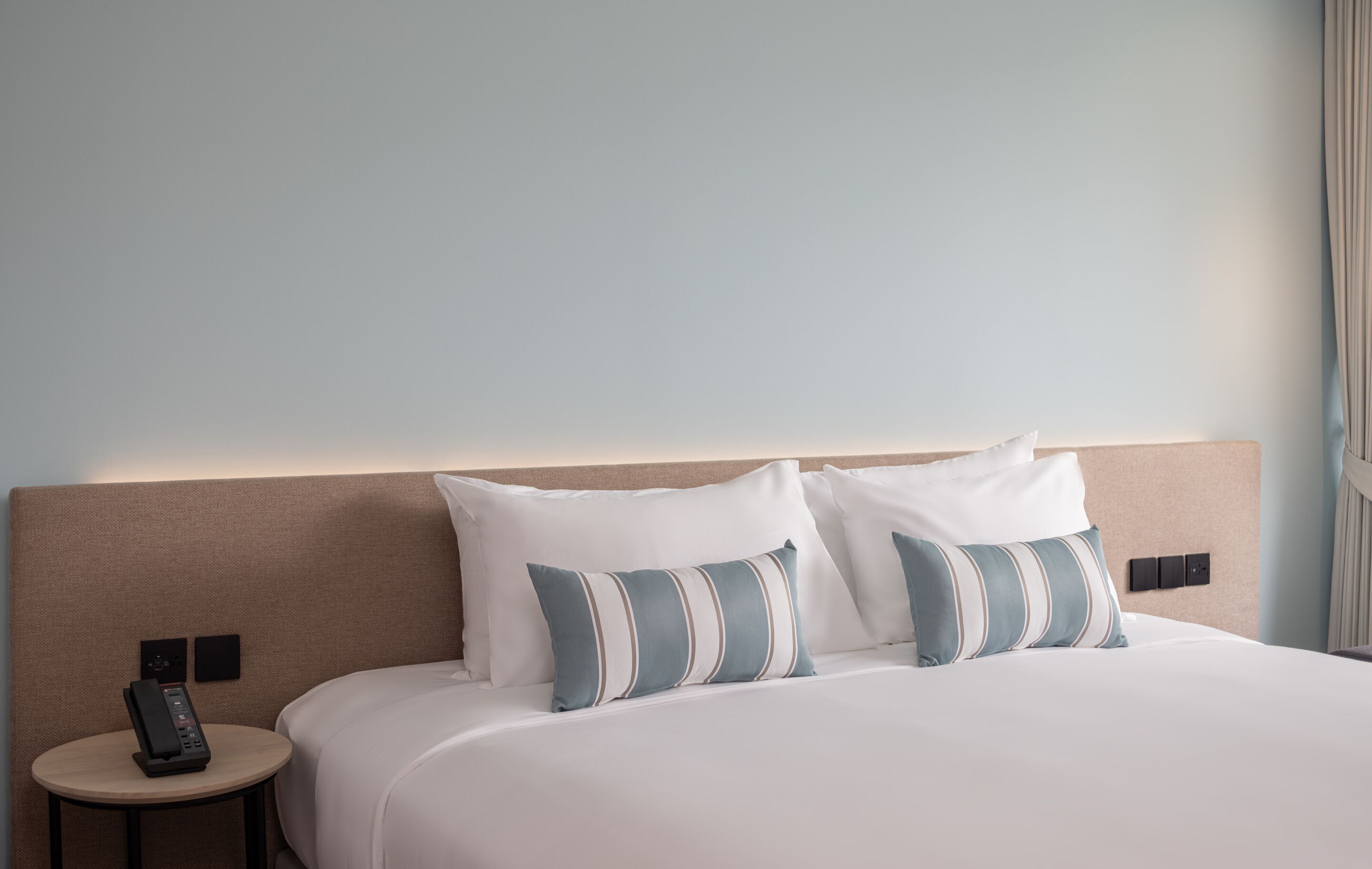 Een hotelkamer met een groot bed, twee kussens, een nachtkastje met een telefoon en een bedieningspaneel aan de muur.