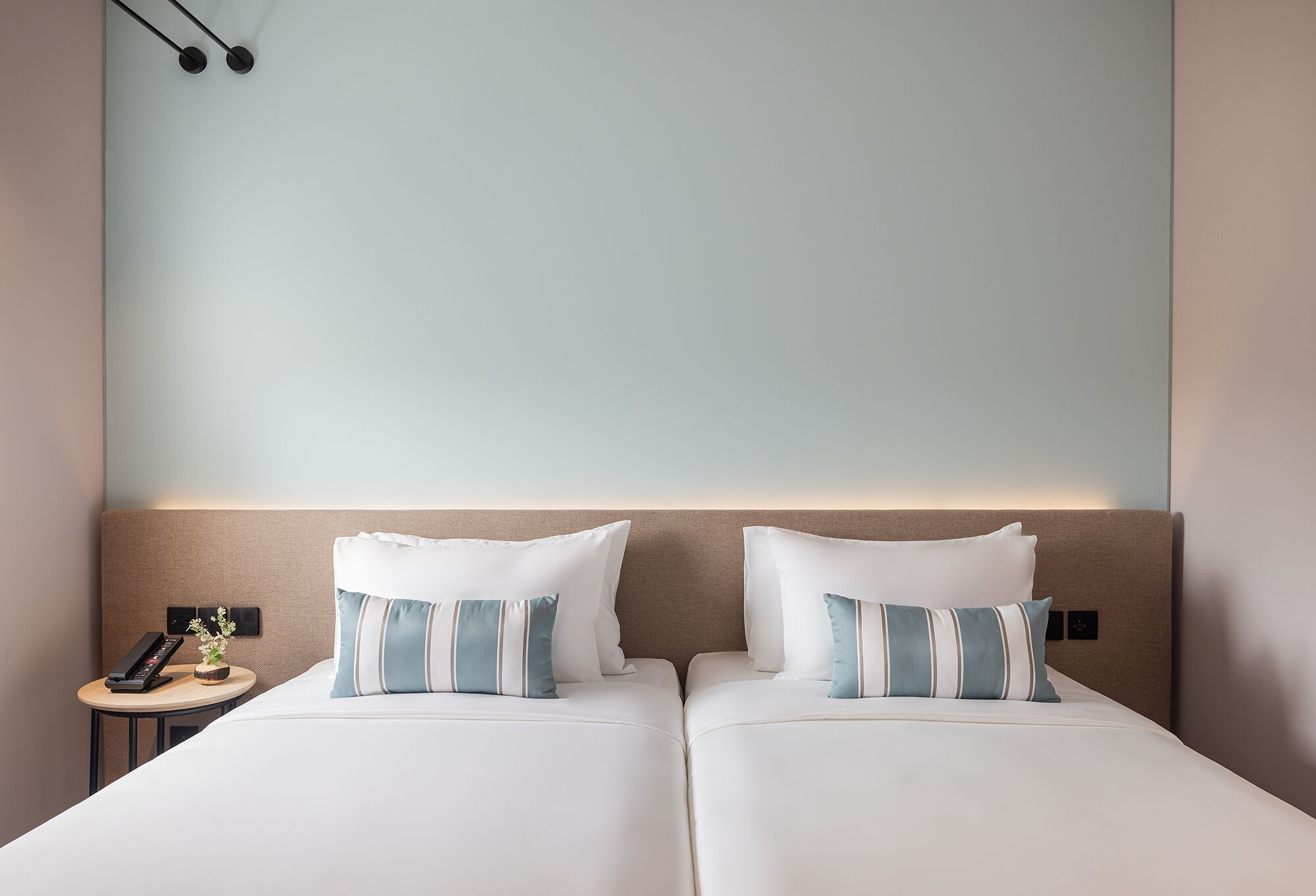 A hotel room with two beds, each with white bedding and striped pillows, a bedside table with a phone and a small plant, and a wall-mounted light fixture.