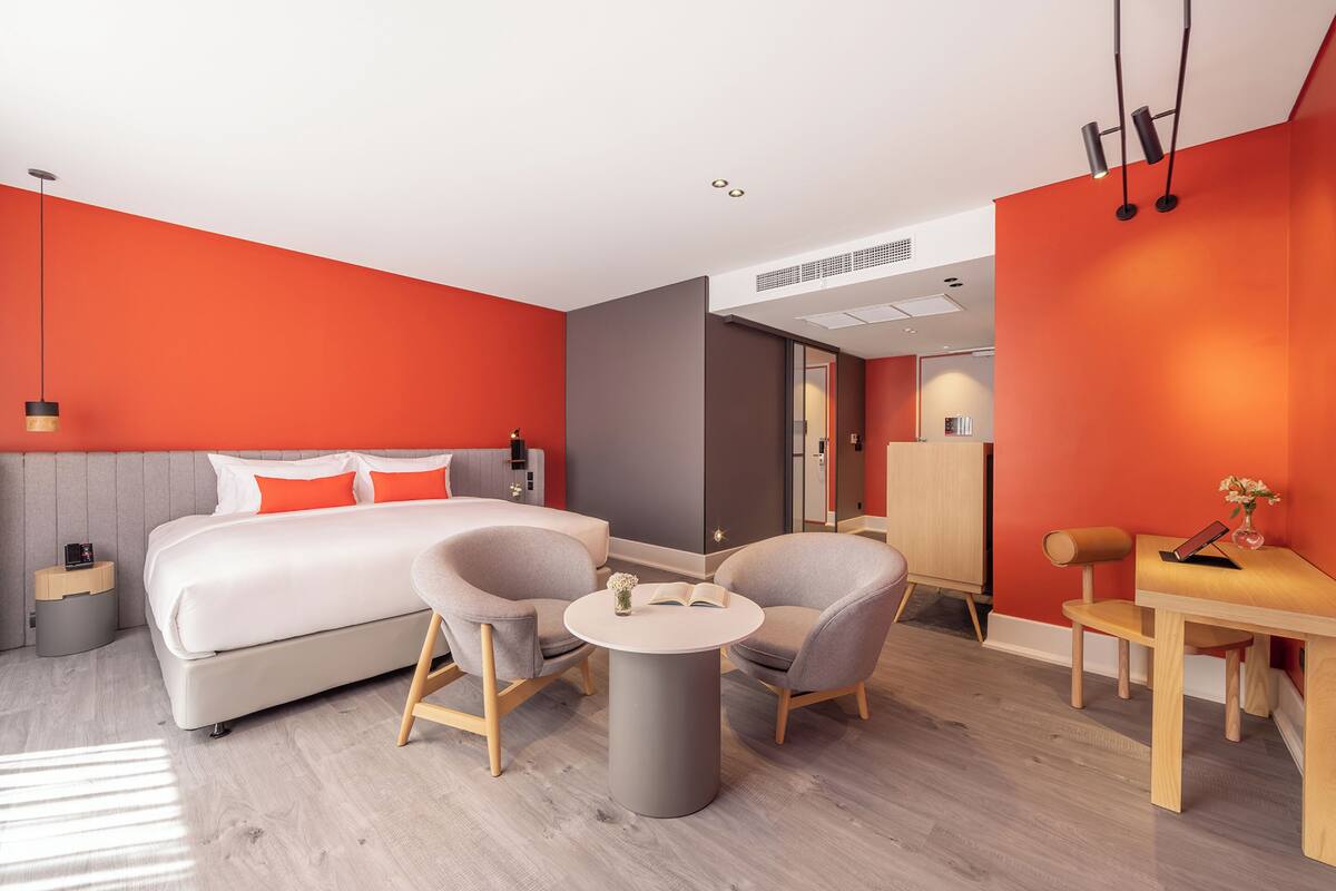 A modern hotel room with a large bed, two armchairs, a small round table, and a desk with a chair.