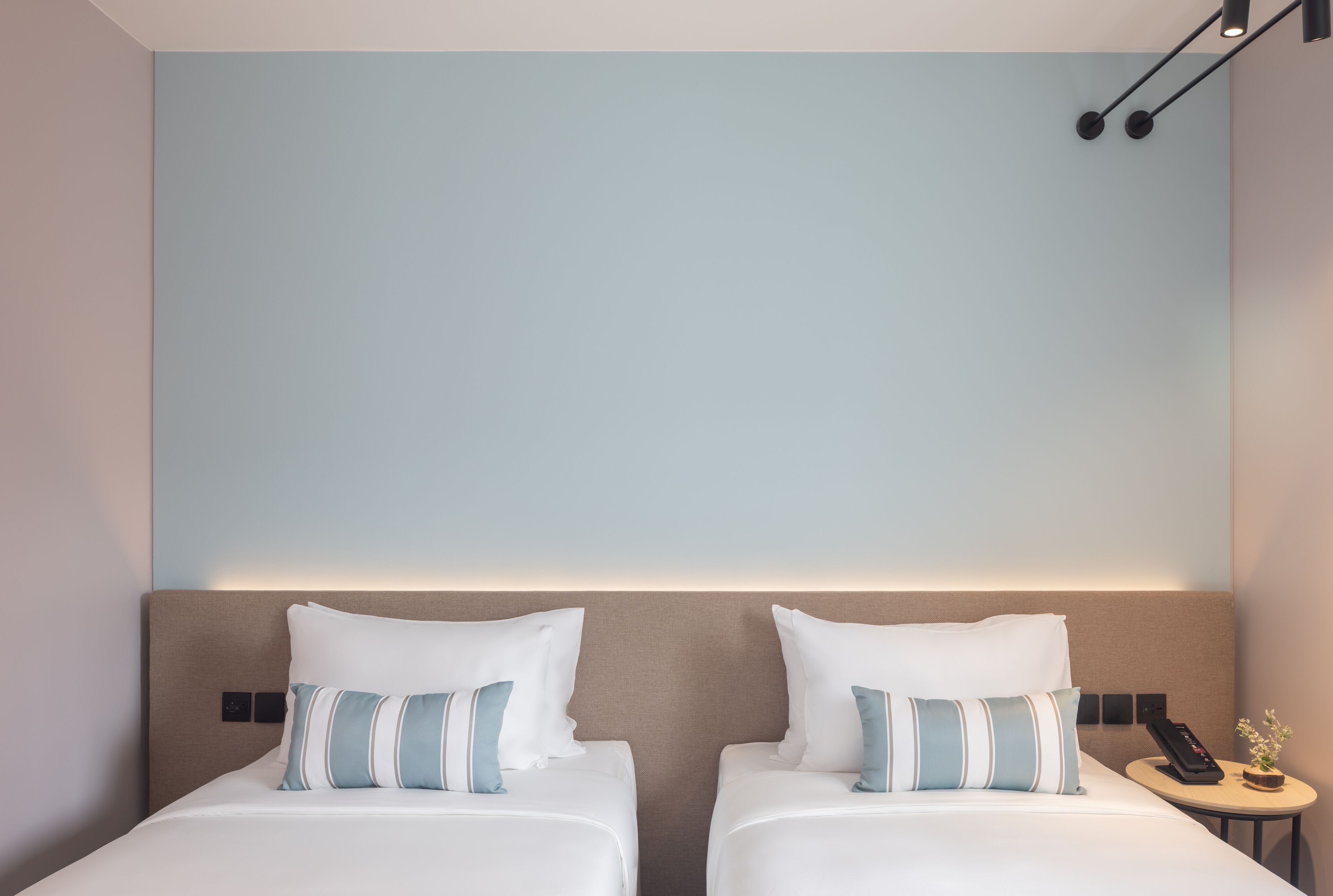 Habitación de hotel con dos camas, cada una con ropa blanca y almohadas a rayas, una mesita de noche pequeña con un teléfono y una planta, y una pared azul claro.