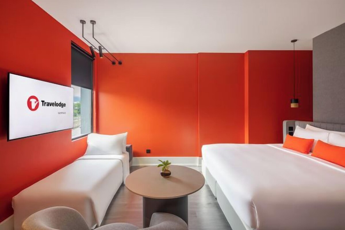A modern hotel room with a red accent wall, a bed, a sofa, a small round table, and a TV displaying the Travelodge logo.