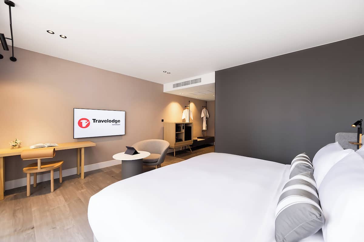 A modern hotel room with a large bed, a desk, a chair, and a TV displaying the Travelodge logo.
