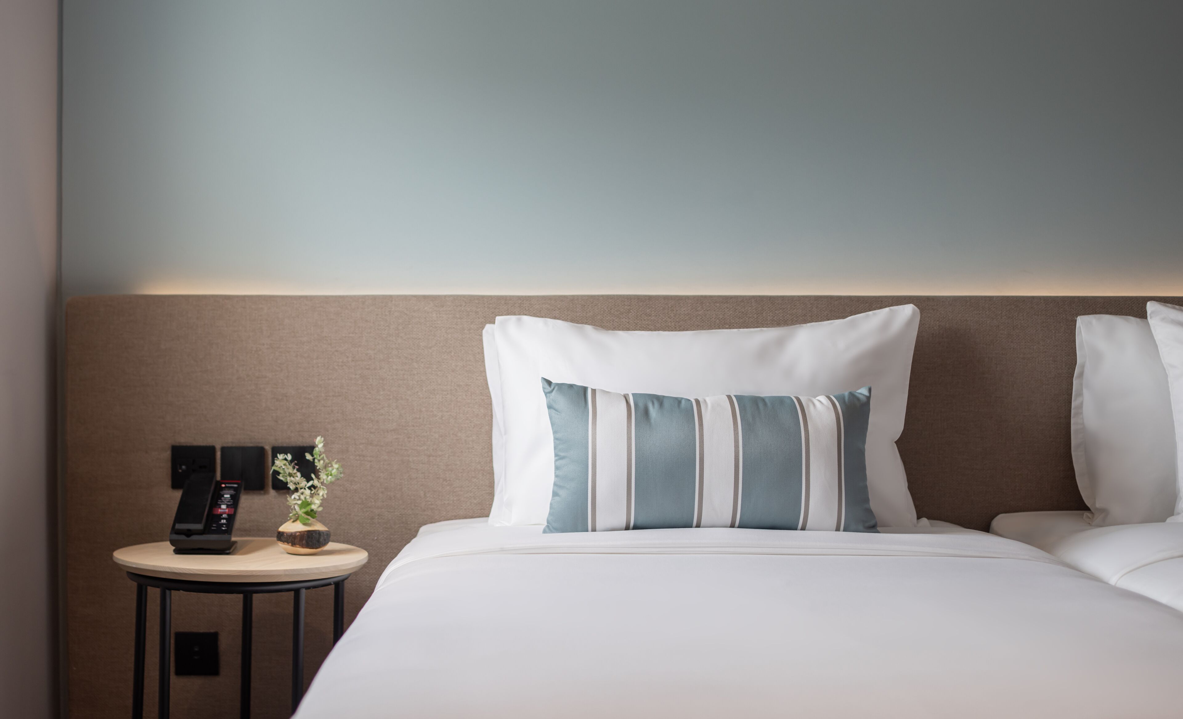 A hotel room with a neatly made bed, a bedside table with a phone and a small plant, and a wall-mounted light fixture.