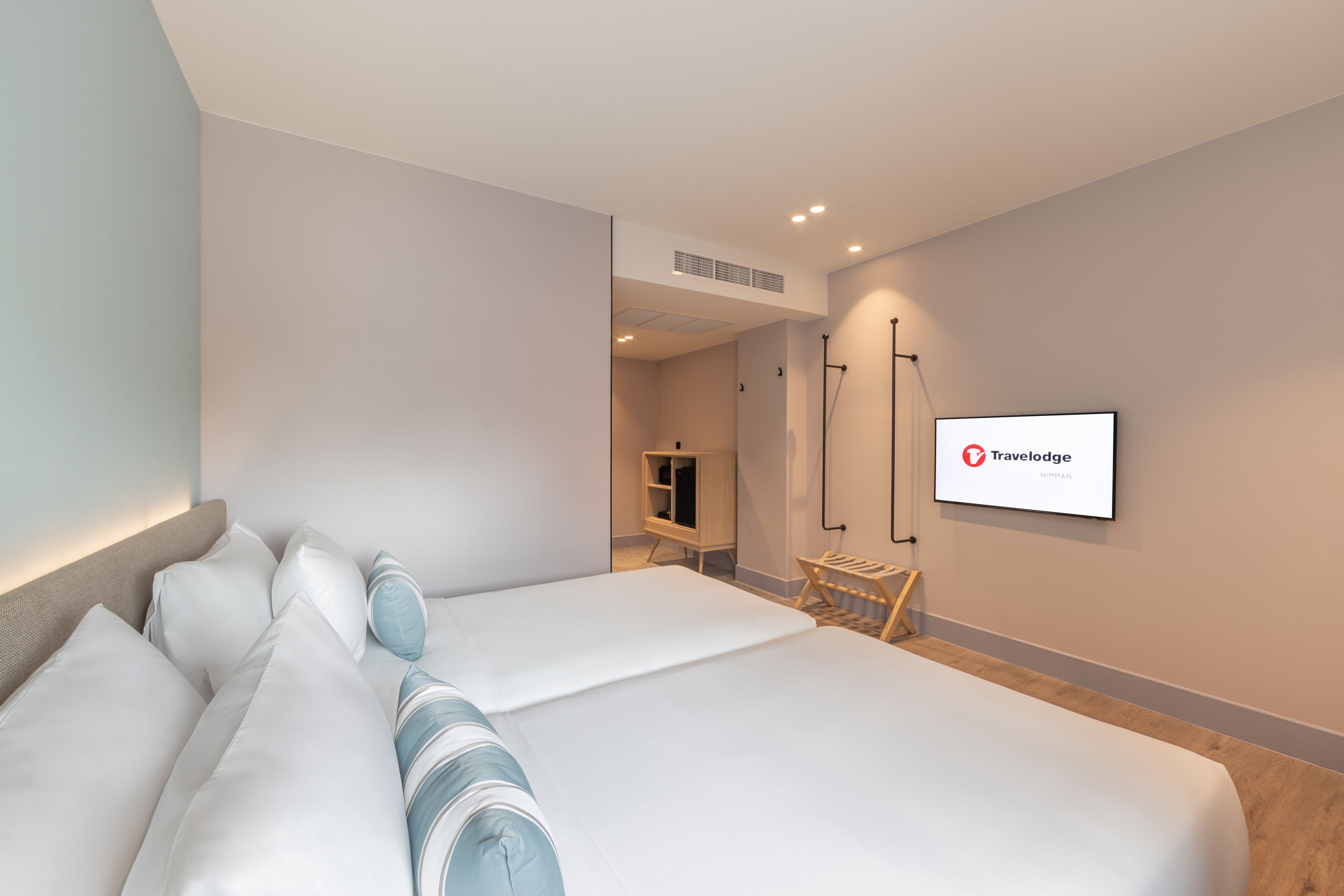 A modern hotel room with a large bed, a desk, and a chair. There is a TV mounted on the wall and a sign with the hotel's logo.