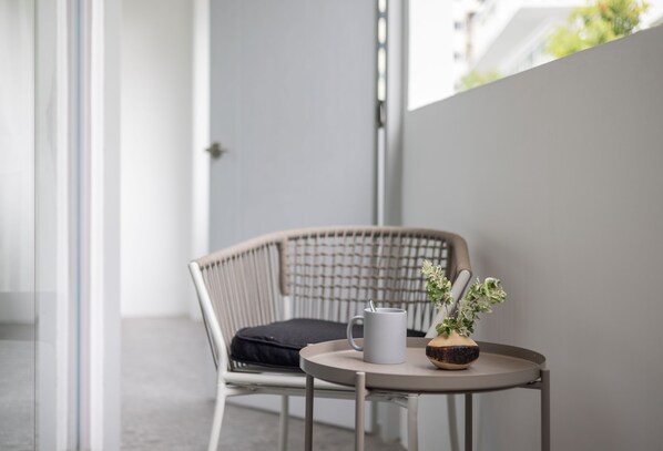 Una silla de mimbre con un cojín, una mesita redonda con una taza blanca y una planta en maceta, y una puerta de cristal que da a un espacio exterior.