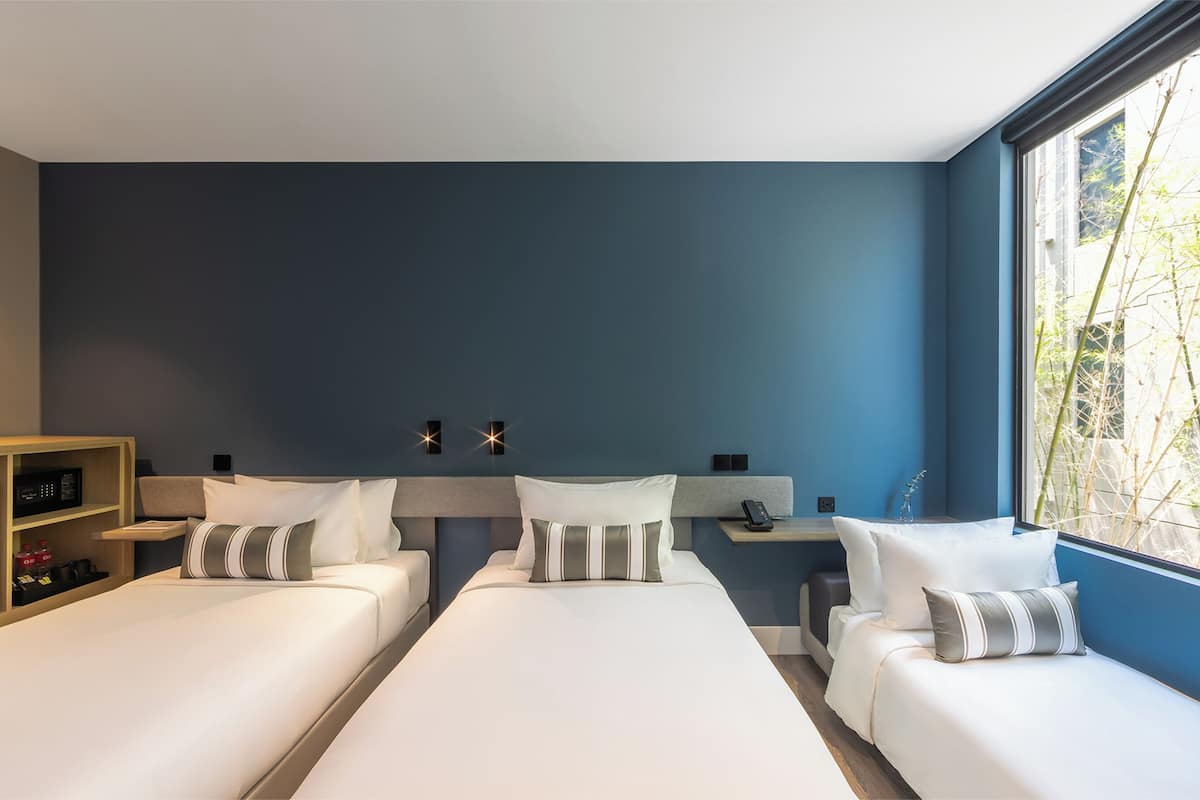 A modern hotel room with two single beds, a small desk, and a large window offering a view of trees.
