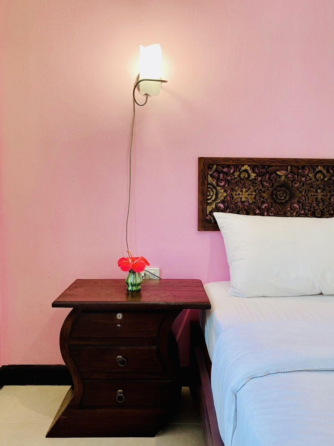 A bedroom with a wooden nightstand, a vase with red flowers, a white pillow, and a headboard with a floral pattern.