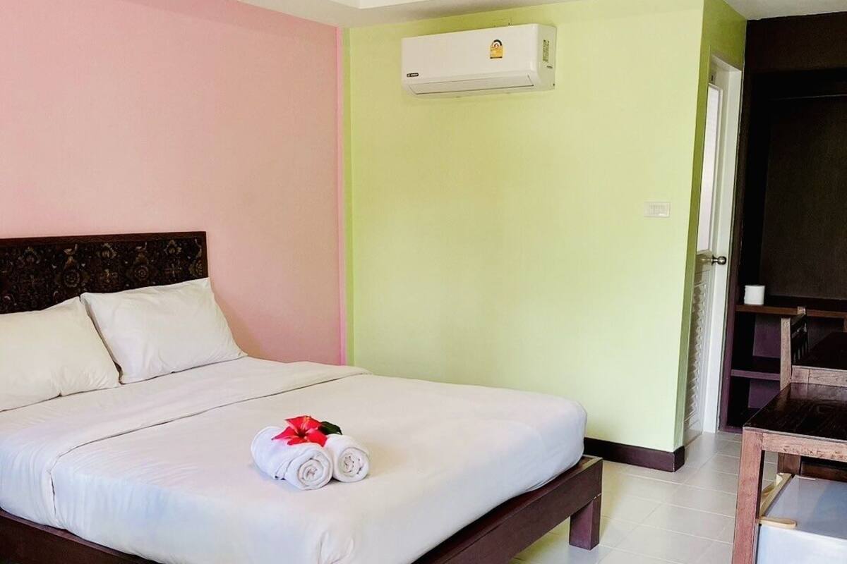 A neatly made bed with white linens and a red flower, a wall-mounted air conditioner, and a wooden nightstand with a white object on it.