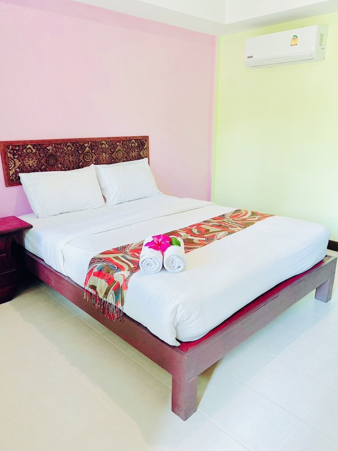 A neatly made bed with a patterned blanket and rolled towels, a wooden headboard, and an air conditioning unit on the wall.