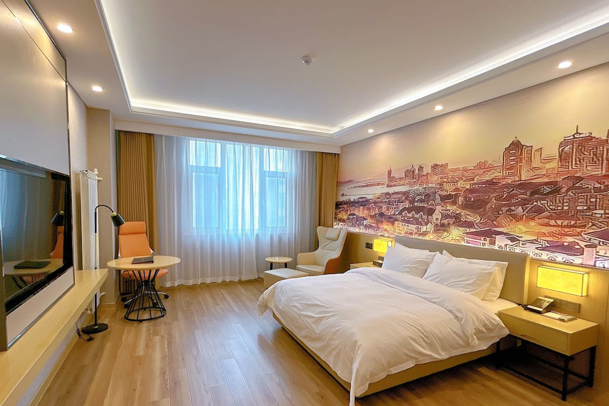 A hotel room with a bed, a desk, a chair, a TV, and a cityscape mural on the wall.