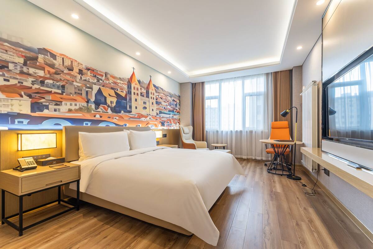 A hotel room with a large bed, a bedside table with a lamp, a desk with a computer, and a large window.
