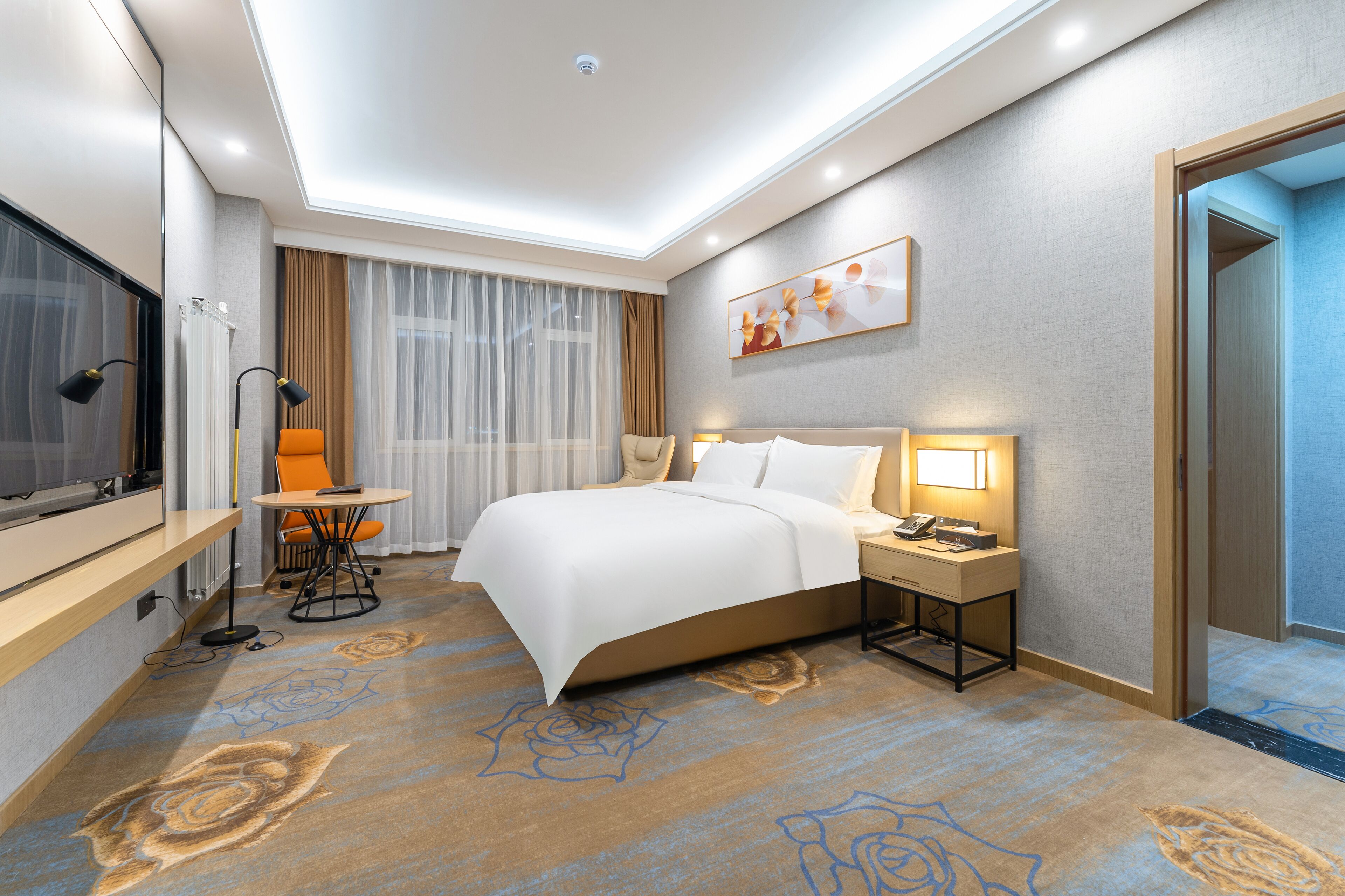 A hotel room with a bed, bedside table, lamp, TV, and a desk with a chair.