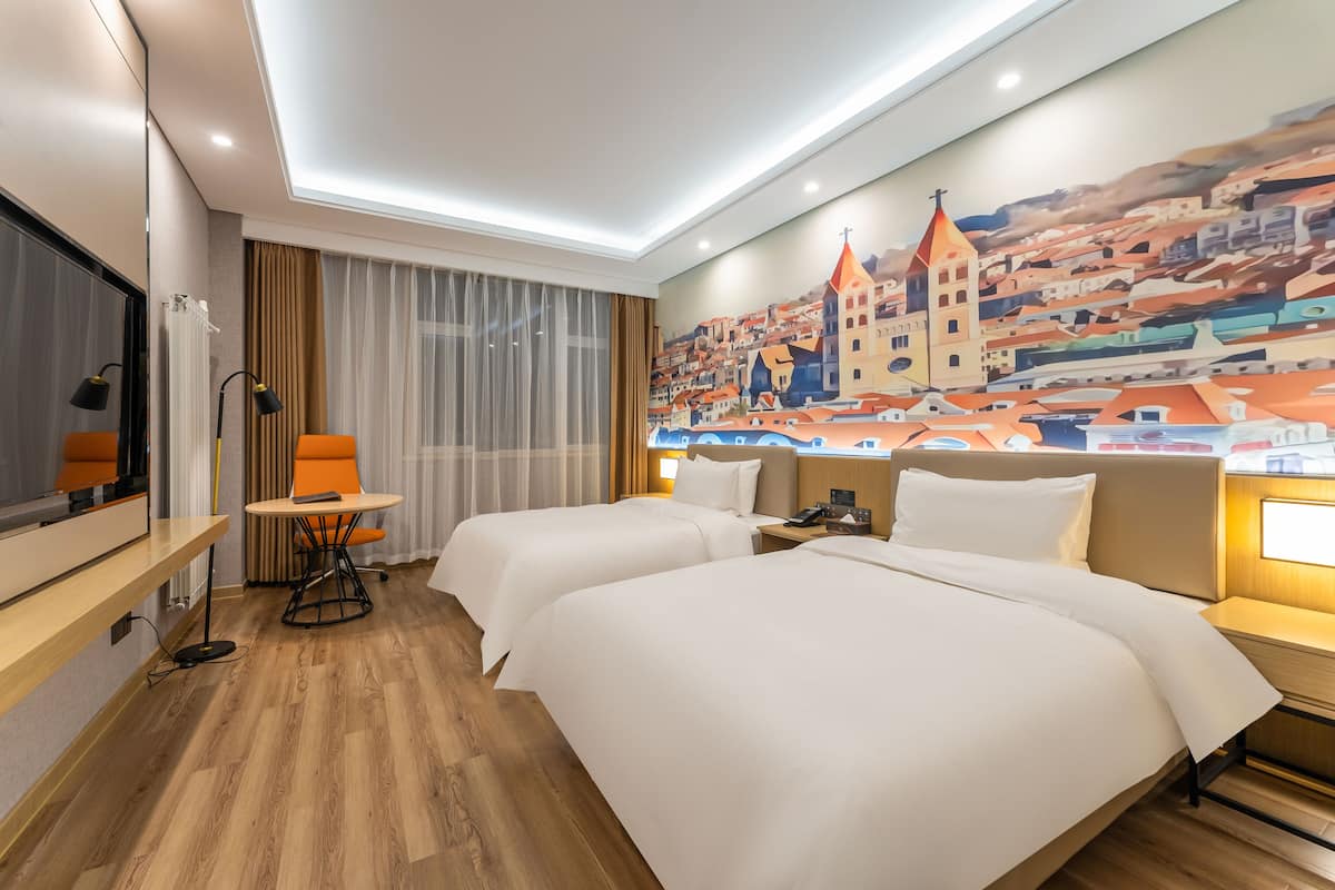 A hotel room with two beds, a desk, a chair, and a cityscape mural on the wall.